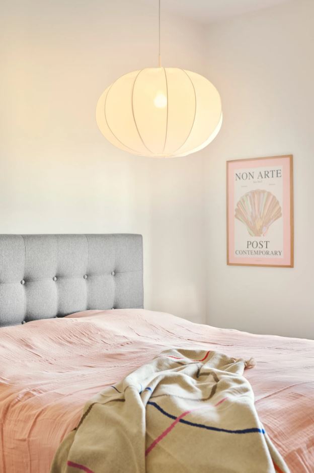 A pendant light with a white, round, fabric shade hanging over a bed in a bedroom setting, with a framed picture above the bed.