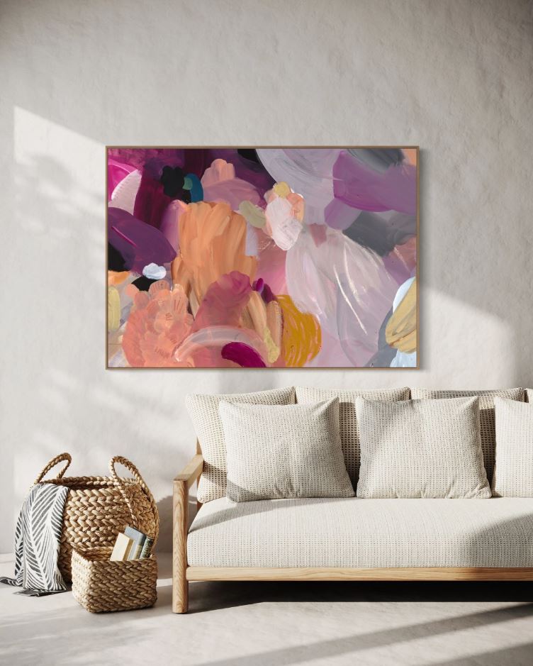 A large abstract painting with predominantly pink and purple hues, featuring textural layers, hanging on a wall above a white sofa in a well-lit room.