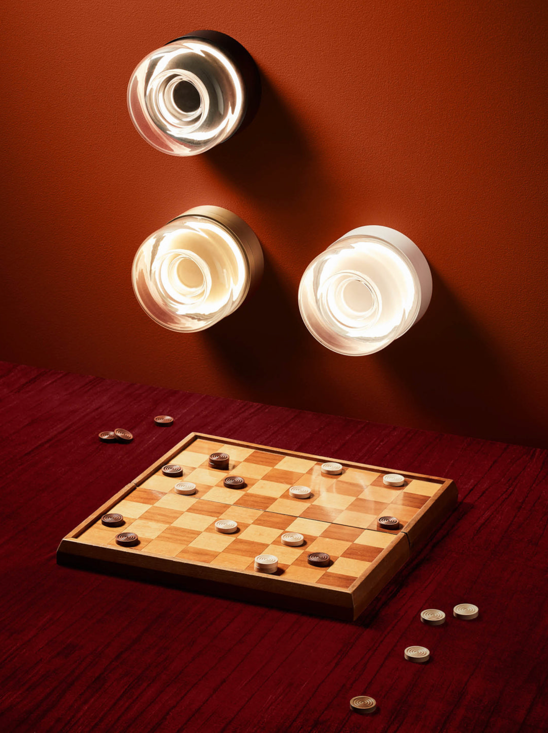 Wooden checkers board with pieces on a red surface, illuminated by three round wall light fixtures.