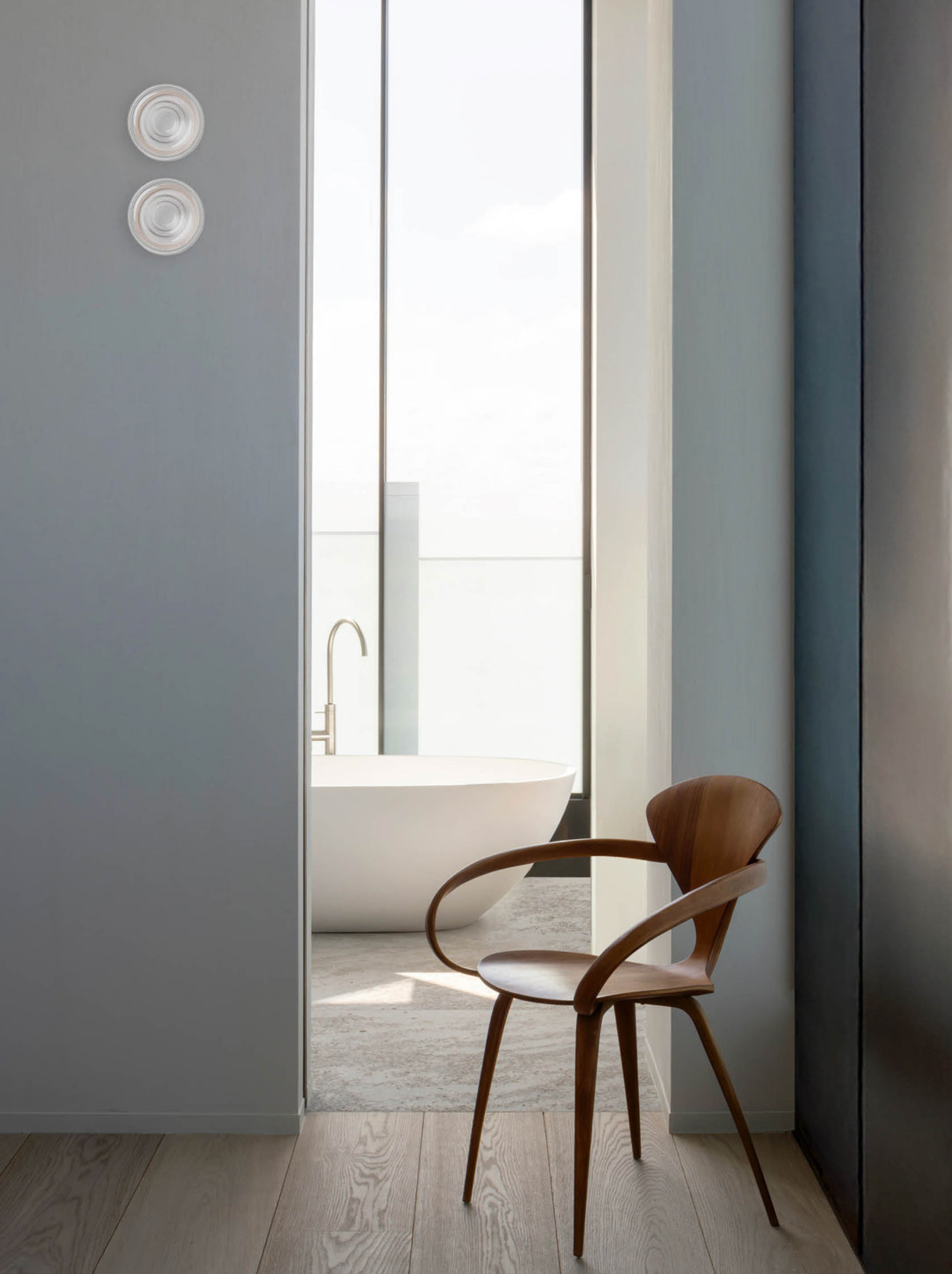 Modern interior with a wooden chair in front of a large window, revealing a bathroom.
