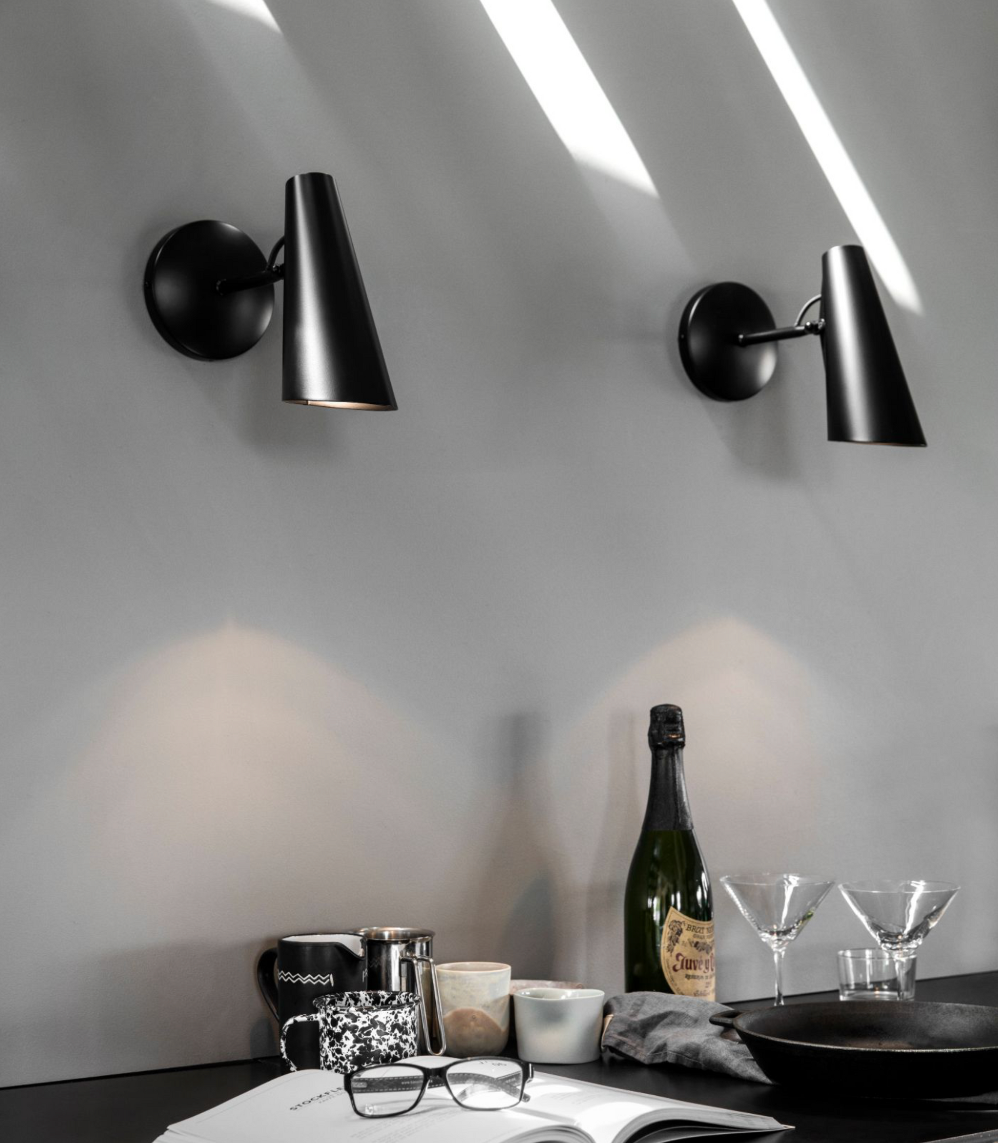 Modern black wall sconces on a white wall with a table setup featuring a bottle, glasses, and books.