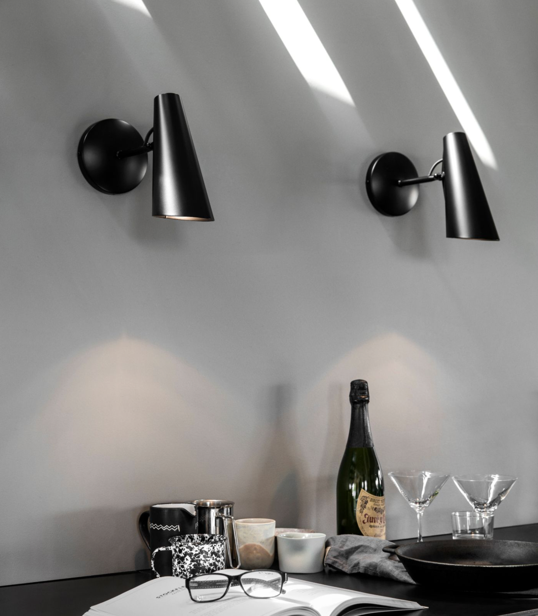 Modern black wall sconces on a white wall with a table setup featuring a bottle, glasses, and books.