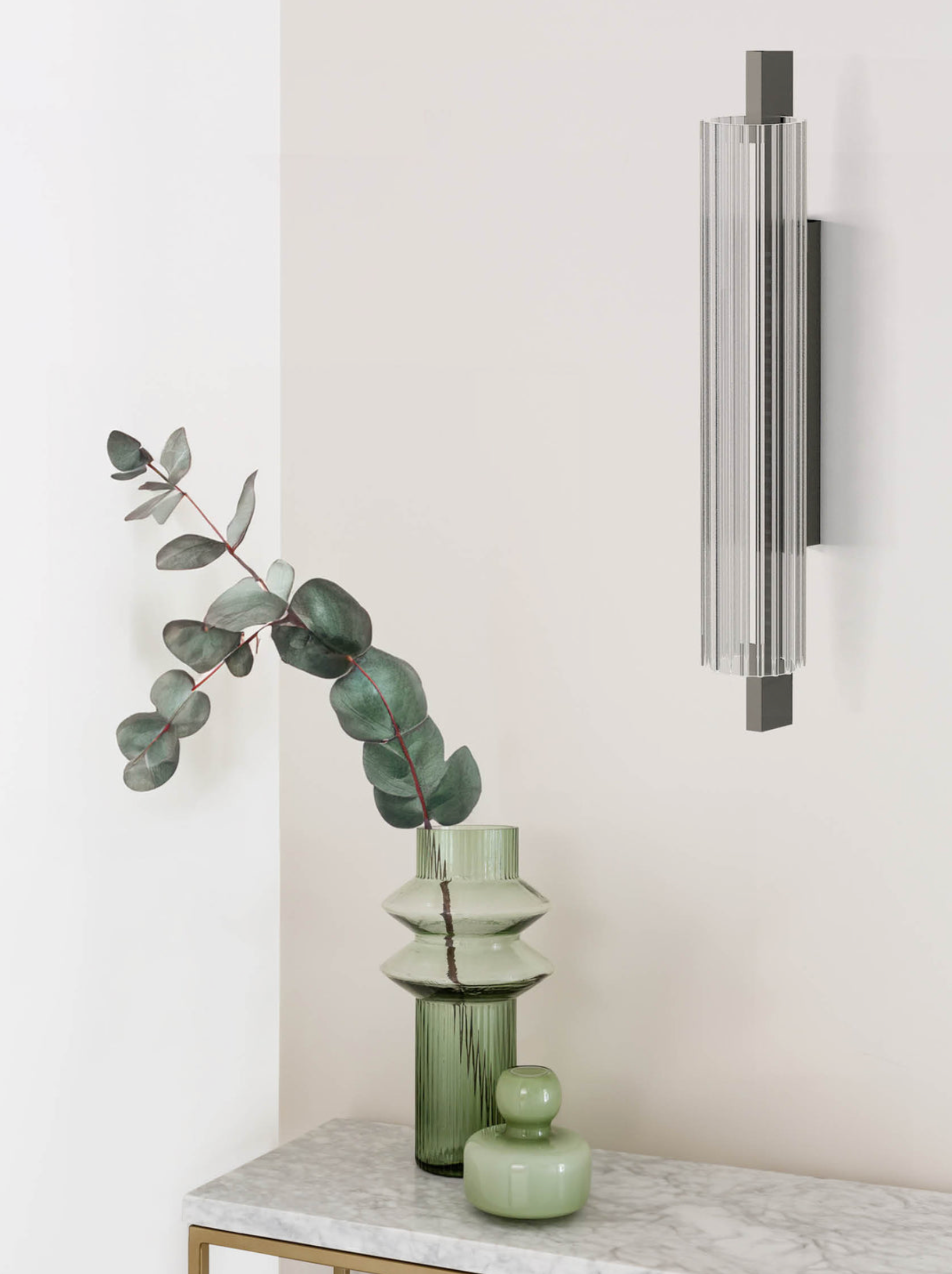 Decorative setup with green vase, eucalyptus branch, and wall-mounted light fixture on a white surface.