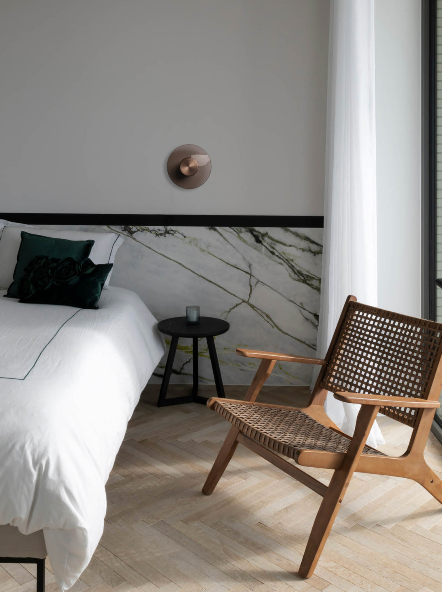 Modern bedroom with a bed, side table, bronze wall light and wicker chair.