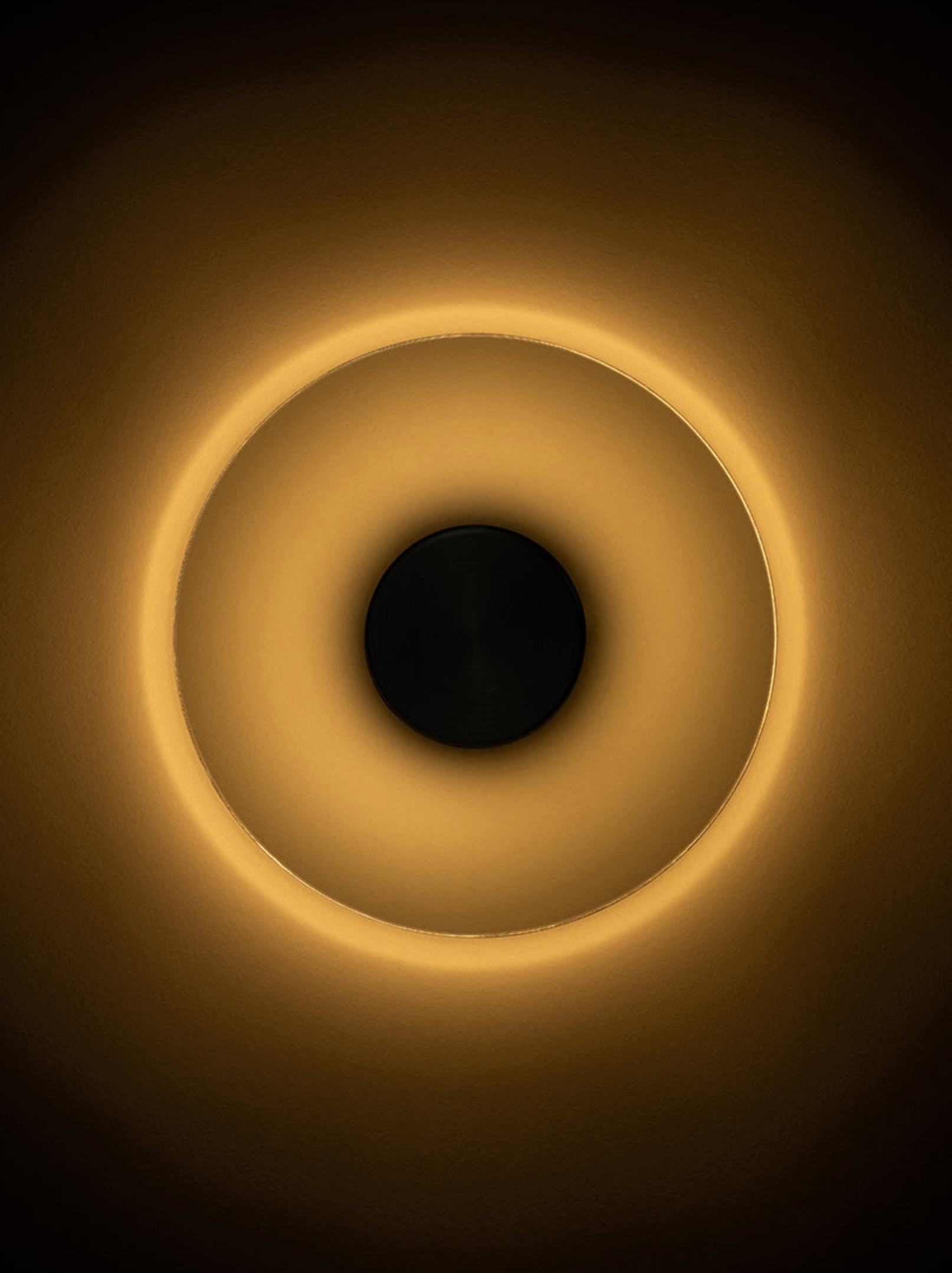 Circular wall light fixture with a dark centre and glowing yellow rim on a dark background
