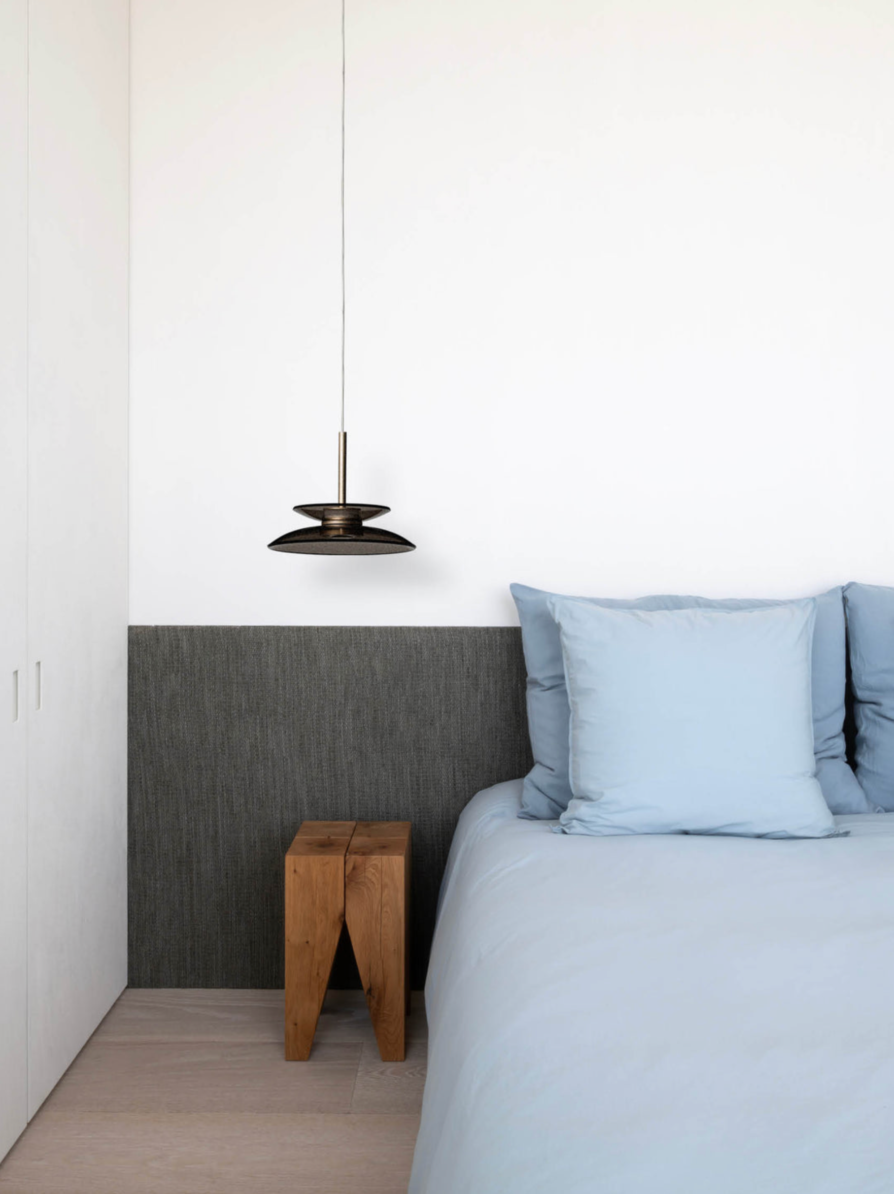 Modern bedroom with grey upholstered headboard, pendant light, wooden side table, and light blue bedding.