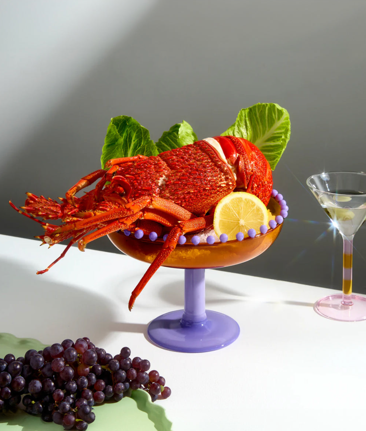 Lobster on a pedestal with lemon and grapes, next to a cocktail glass.