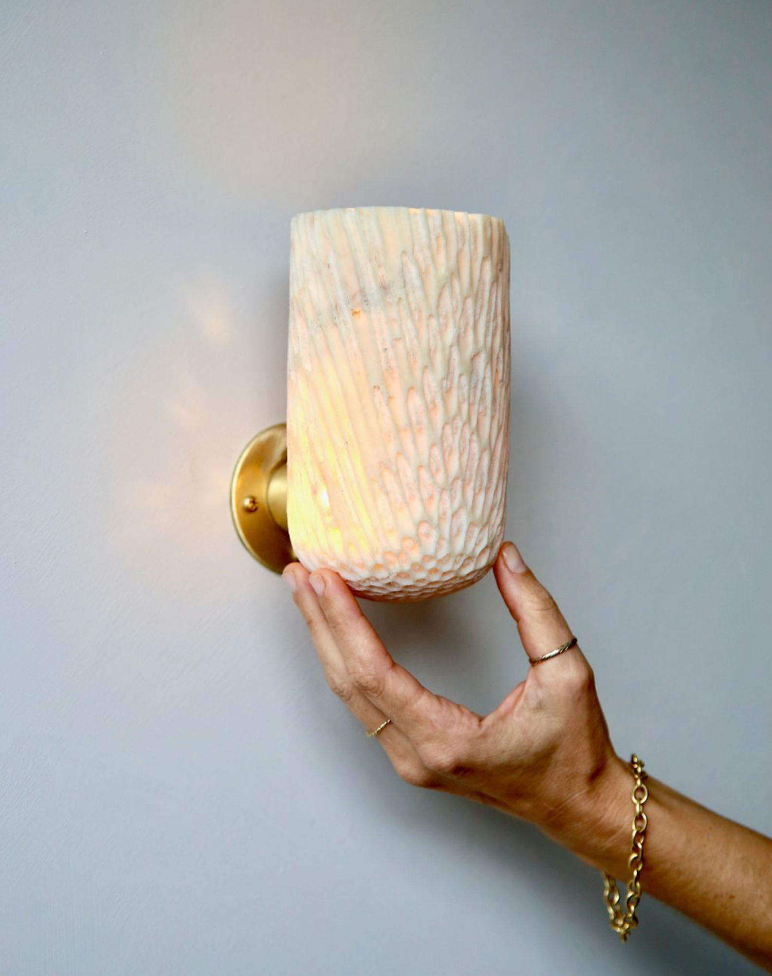 Hand adjusting a wall-mounted light fixture with a textured lampshade against a plain background
