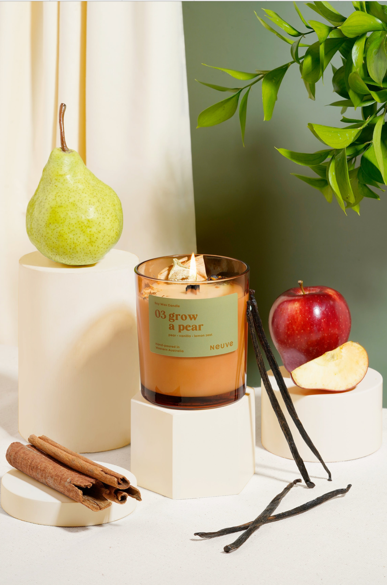 Candle with '03 grow a pear' label surrounded by pears, apples, vanilla beans, and cinnamon sticks on a white surface.