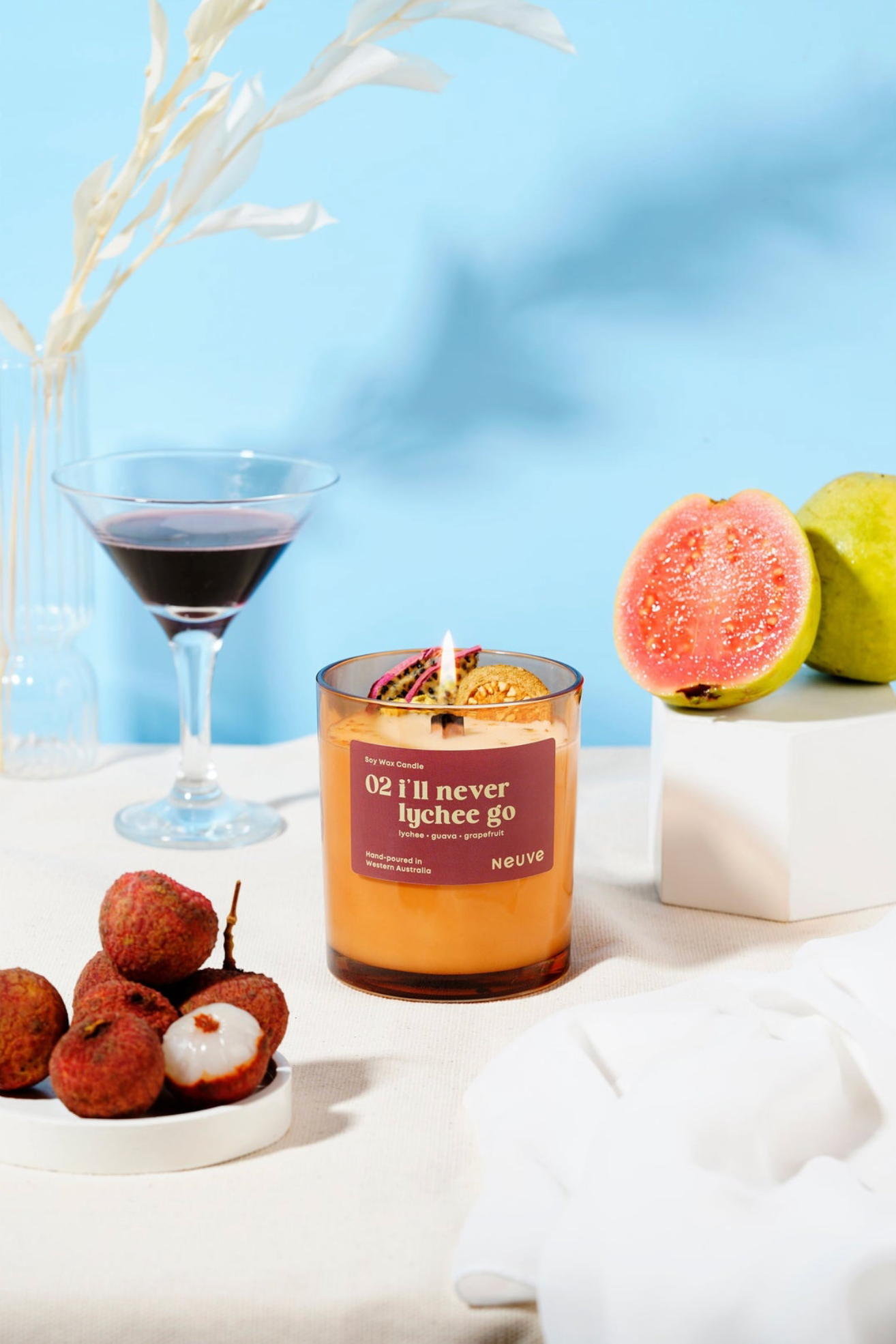 Candle with a humorous label next to fruit and a glass on a light surface with a blue gradient background
