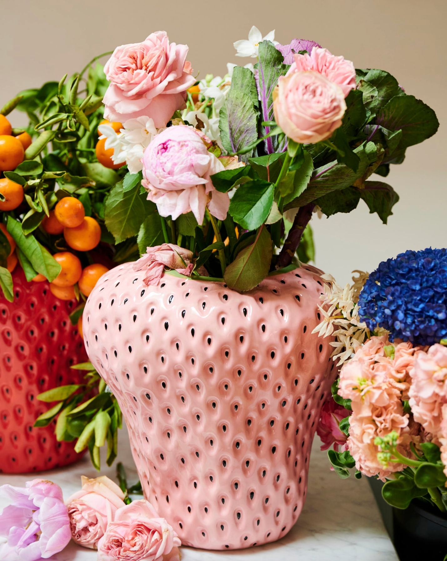 Pink textured vase with flowers surrounded by other floral arrangements on a light surface.