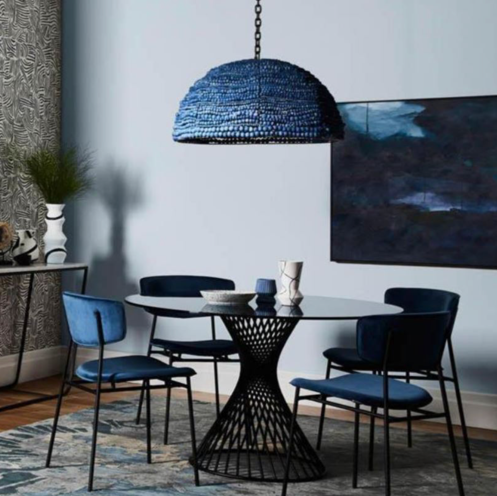 Dining room with a black wireframe table and blue chairs, featuring a blue clay bead pendant light and abstract painting.