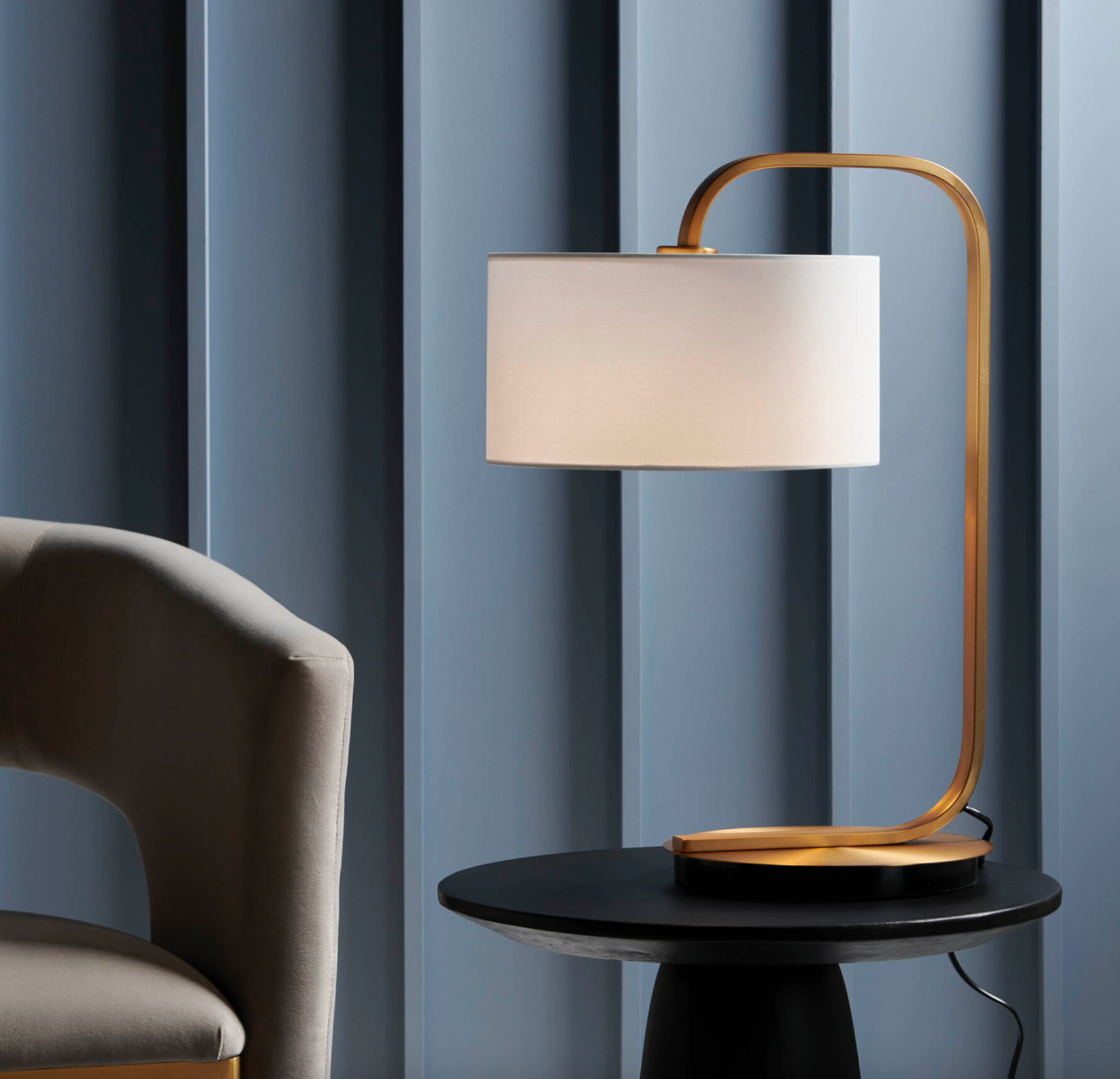 Modern table lamp with gold base and white shade on a black table against a blue curtain background