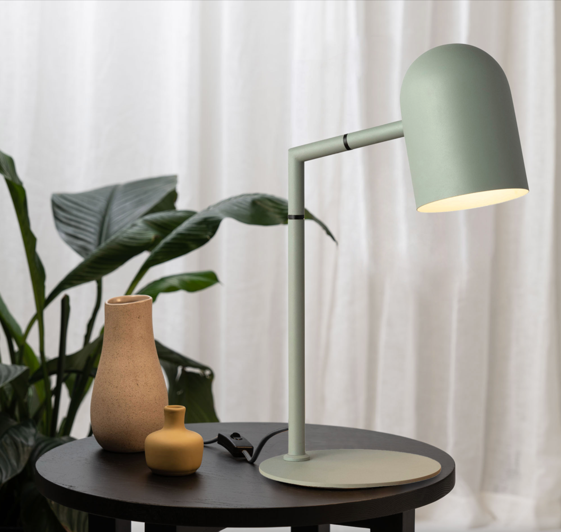 Green desk lamp on a round dark table with a plant and vases in the background