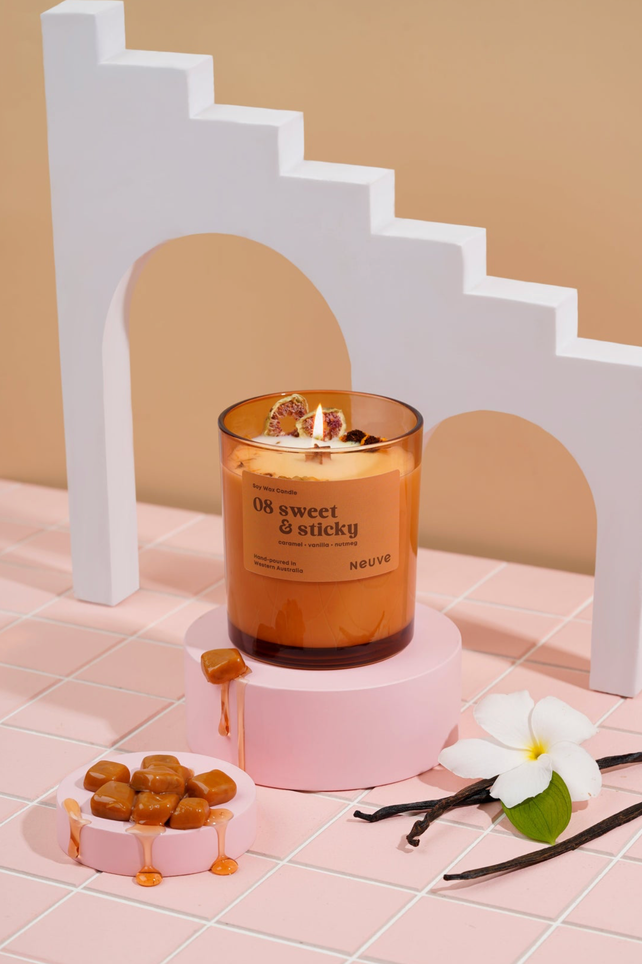 Candle labeled 'Sweet & Sticky' on a pink stand with vanilla beans and flowers on a tiled floor.
