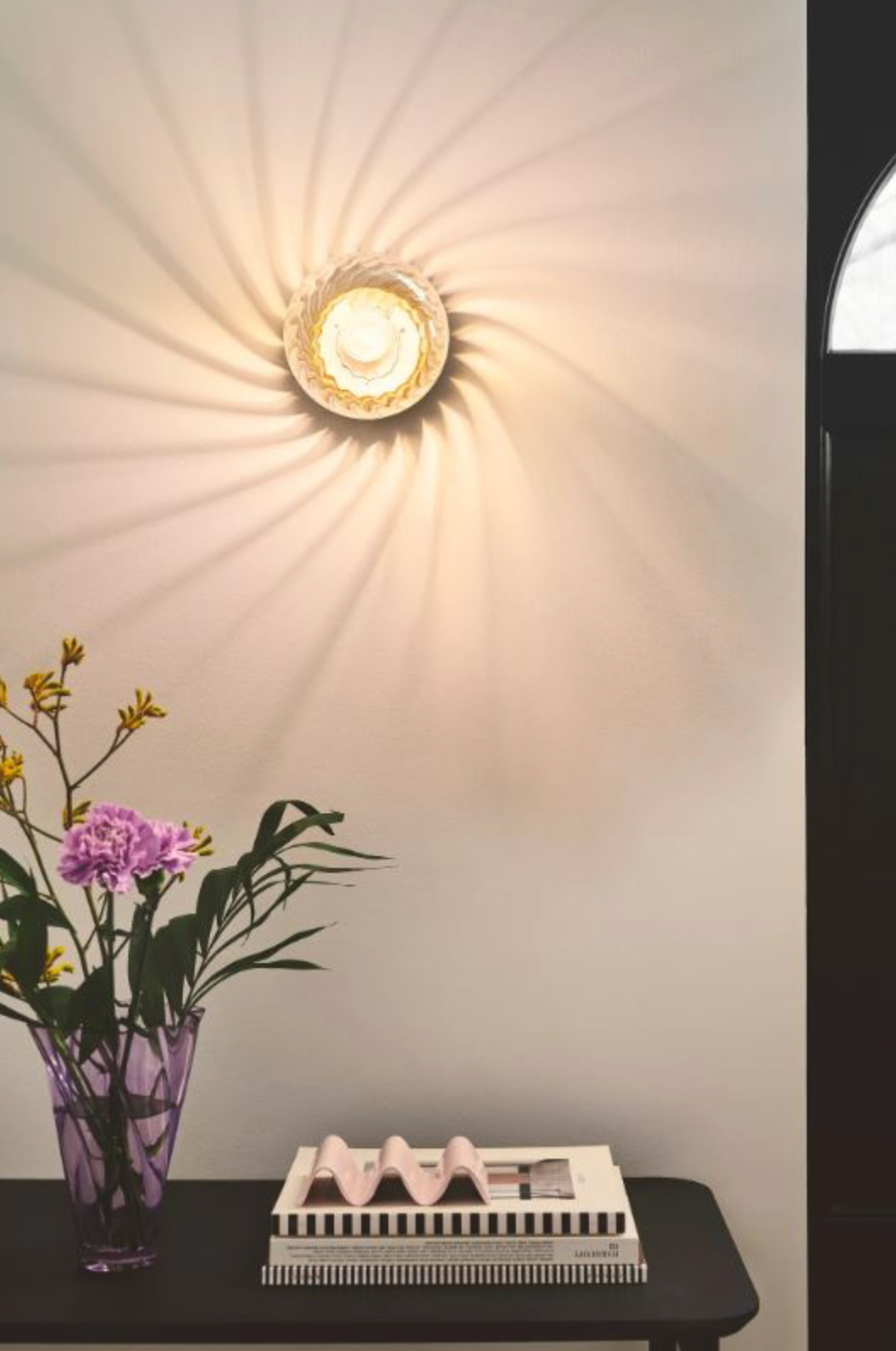 Wall-mounted light fixture casting a warm glow on a wall with a vase of flowers and a magazine on a table below.