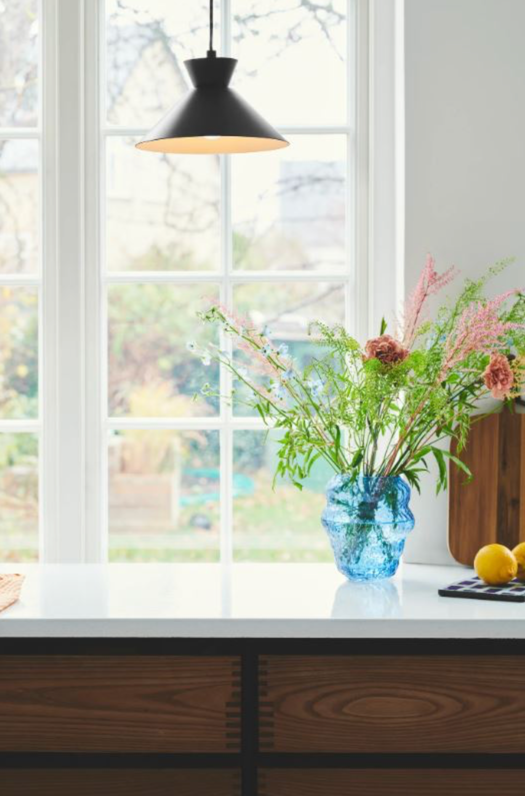 Kitchen counter with a blue vase of flowers, black pendant light, and window view.