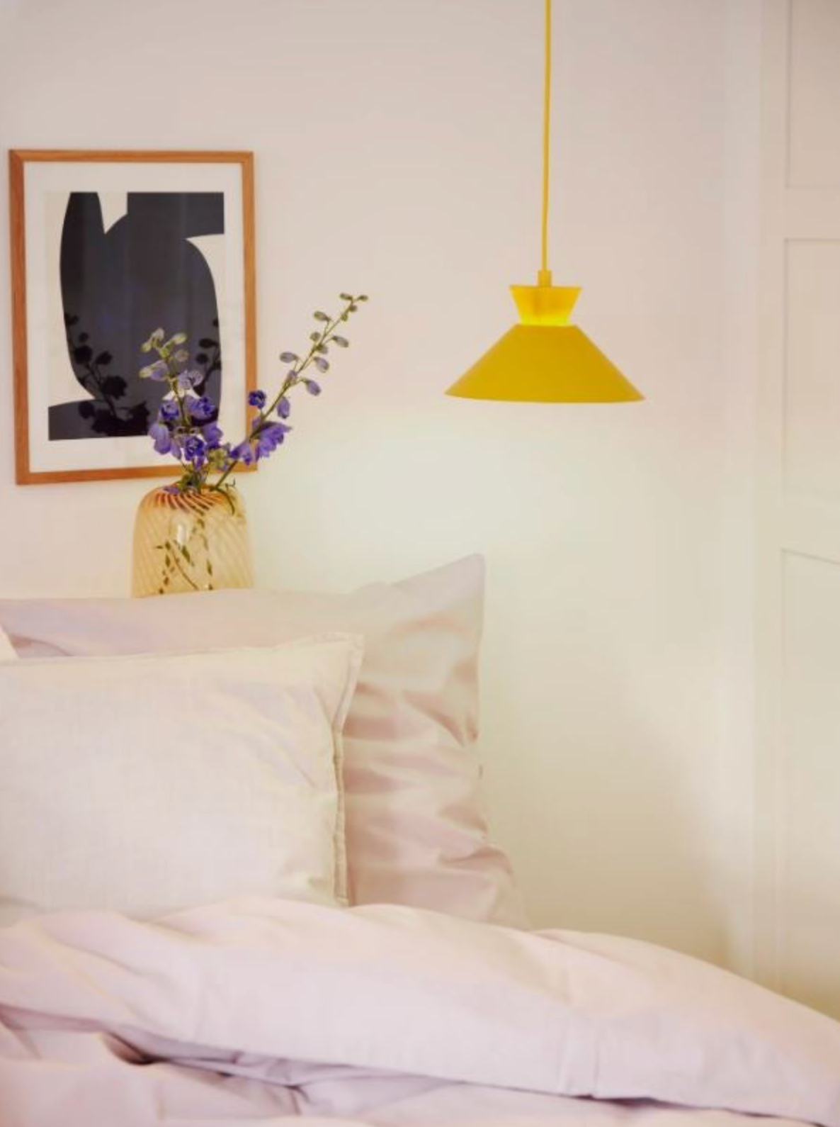 Yellow pendant light above a bed with a vase of flowers and abstract art on the wall.