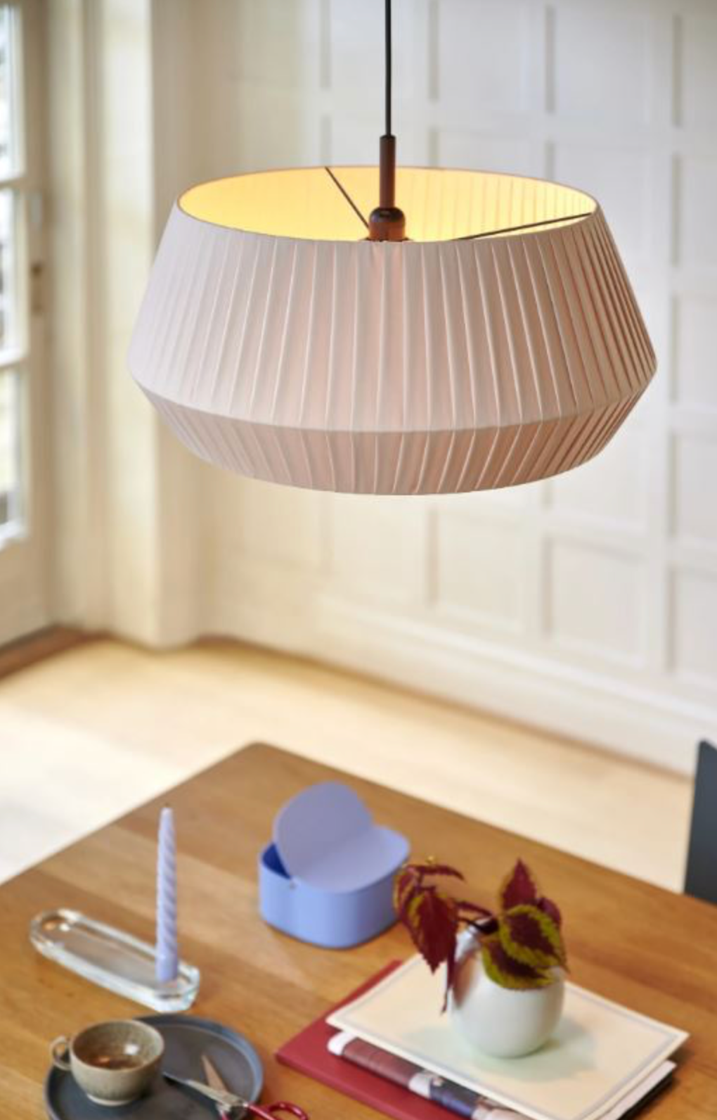 Modern pleated fabric pendant lamp hanging above a dining table with a cup and saucer and decorative items.