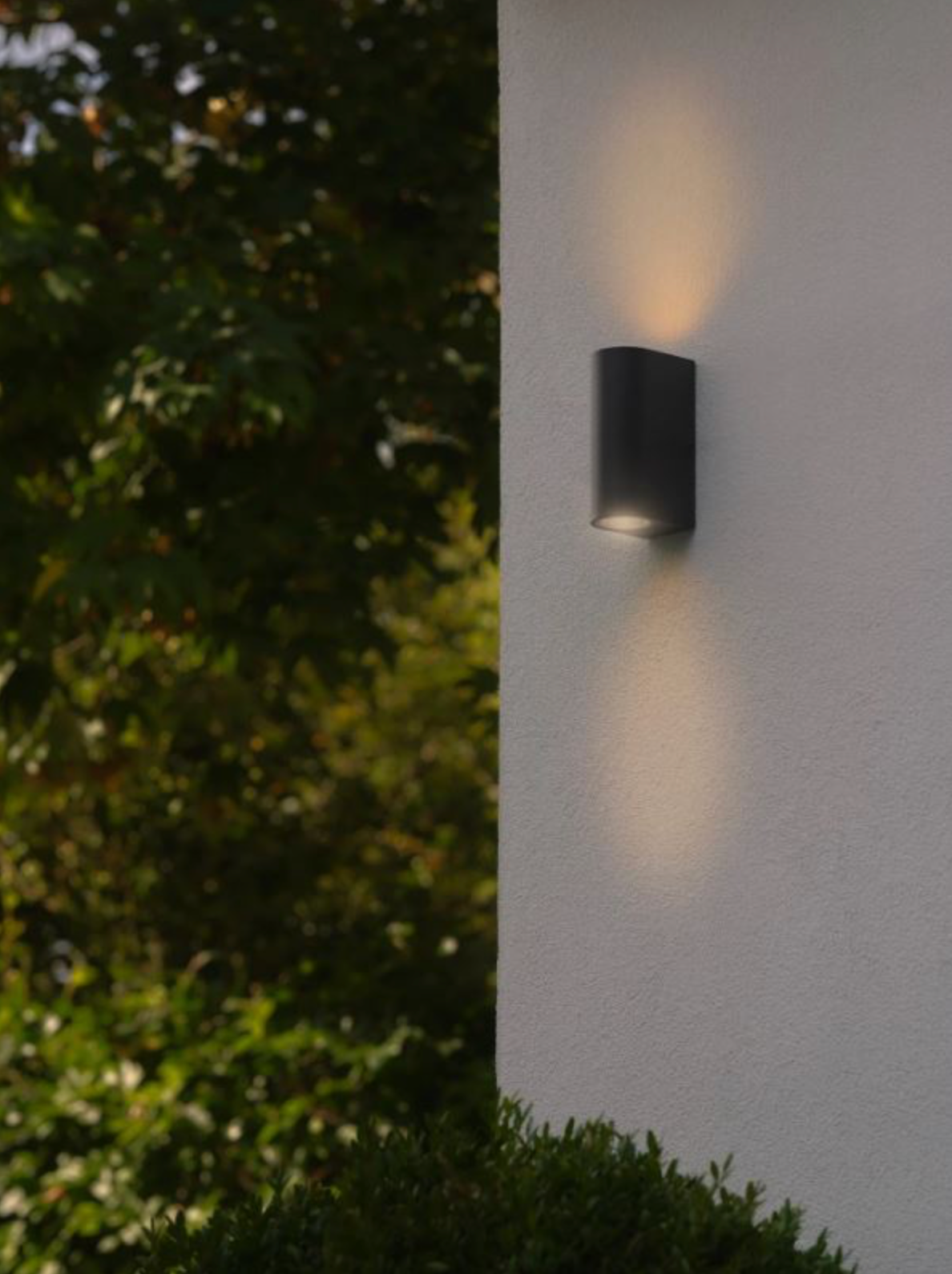 Black outdoor wall light fixture on a white wall with greenery in the background