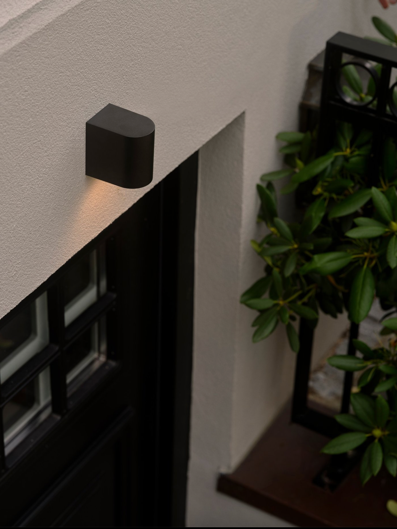 Black outdoor wall light fixture on a building exterior with a plant in the background.