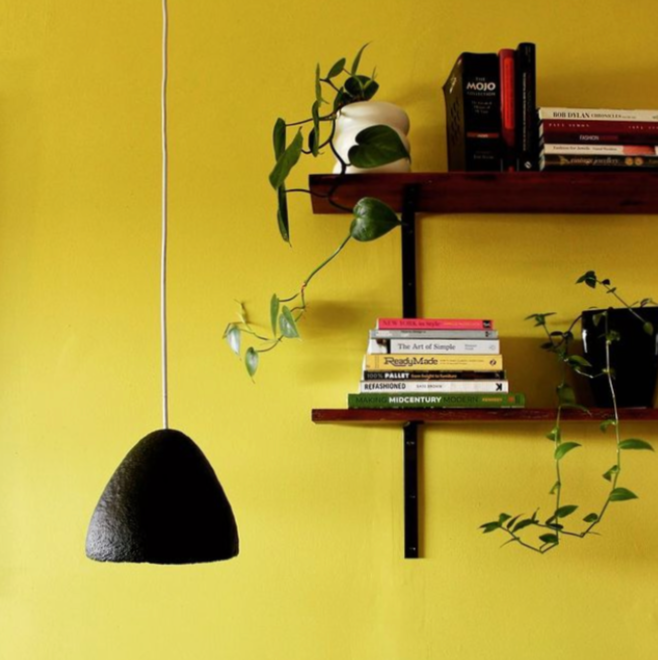 Black textured pendant light hanging above a wooden shelf with books and plants against a yellow wall.