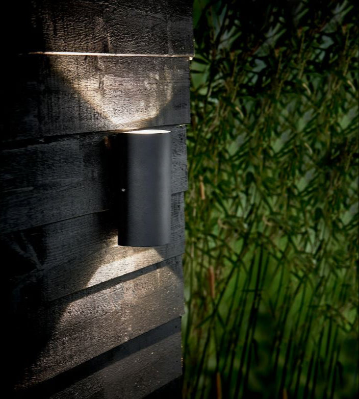 Wall-mounted outdoor light fixture on a wooden wall with greenery in the background