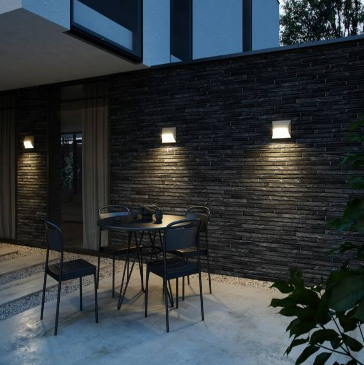 Outdoor dining area with modern chairs and table illuminated by out door wall lights against a dark brick wall.