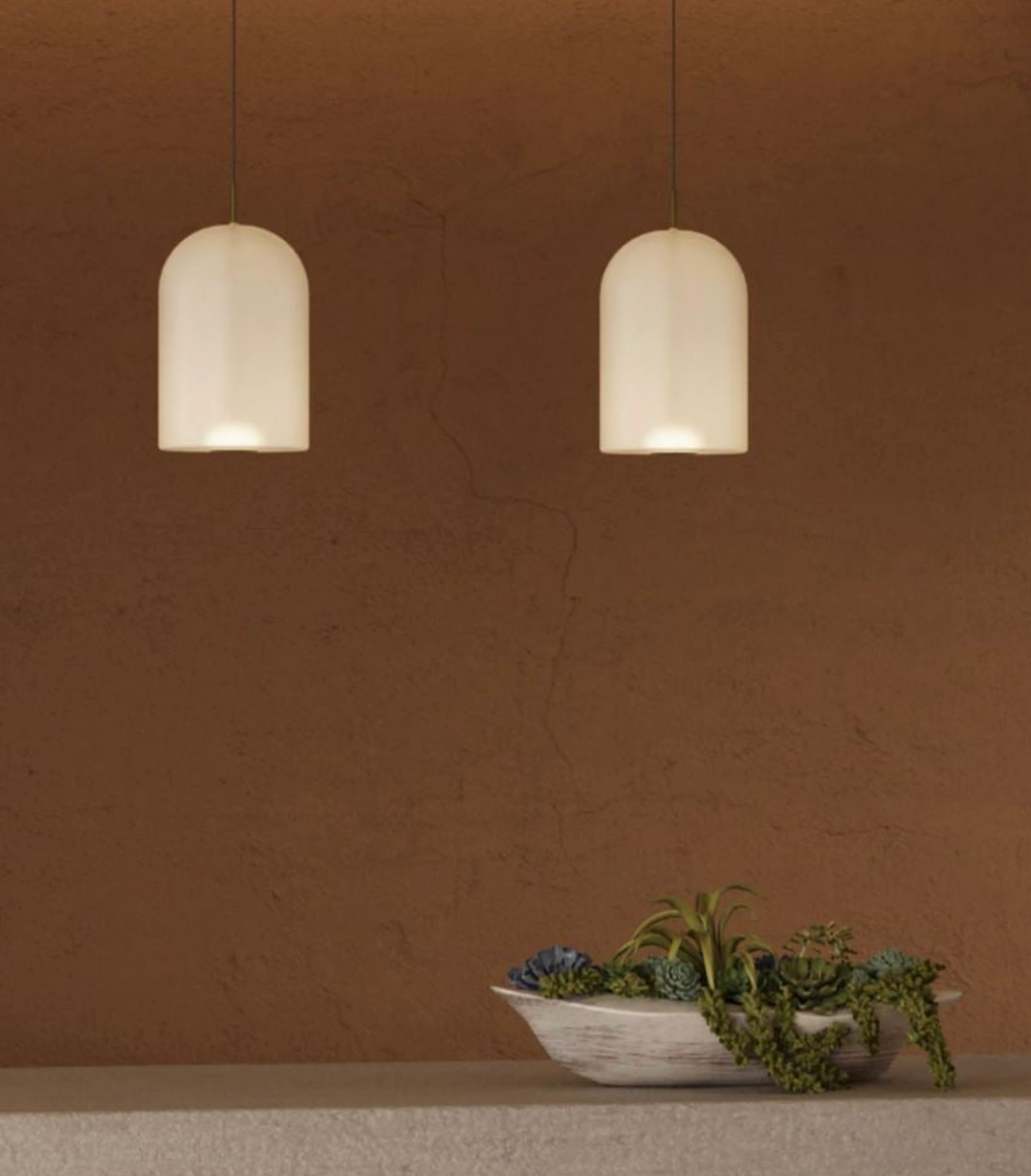 Two pendant lights hanging against a brown wall with a bowl of plants on a surface.