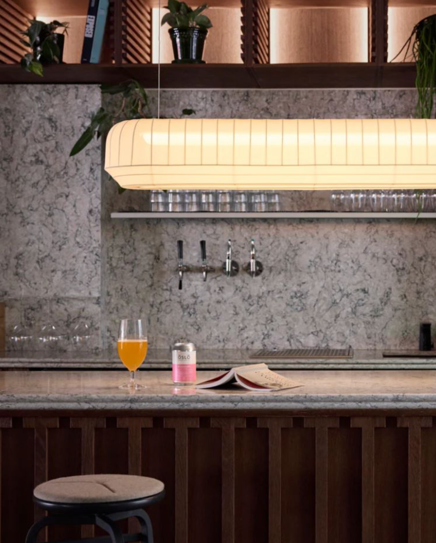 Bar counter with a decorative linear pendant light, glass of beer and a can, set against a marble backsplash.