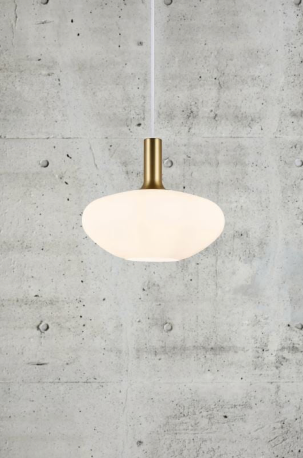 Pendant light with white shade and brass fixture against a concrete wall.