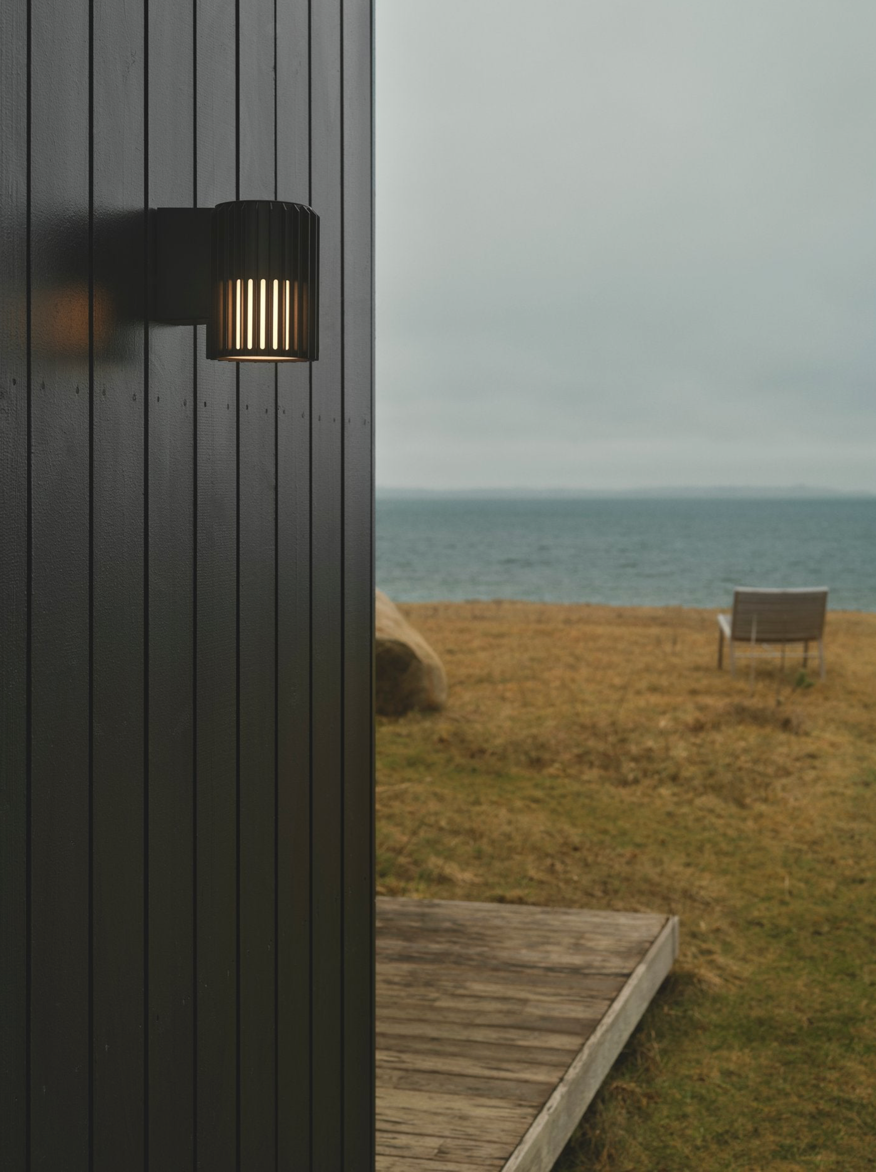 Outdoor wall light fixture on a black wooden wall with a bench and grassy area in the background.