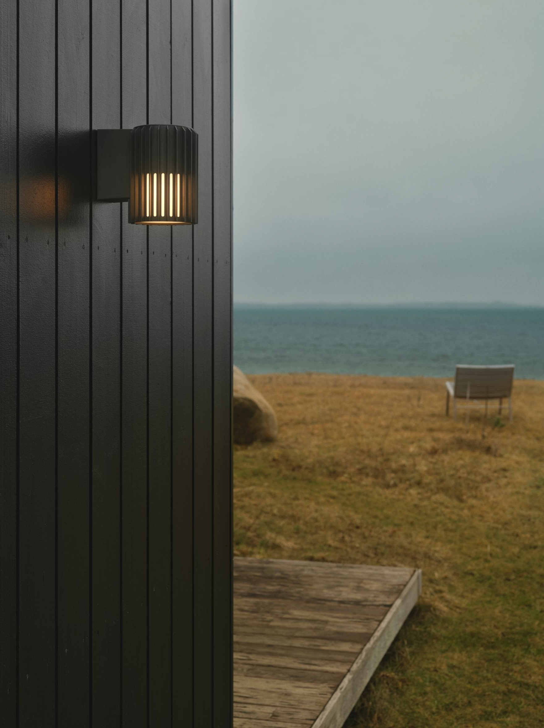 Outdoor wall light fixture on a wooden building with a grassy area and bench overlooking water.