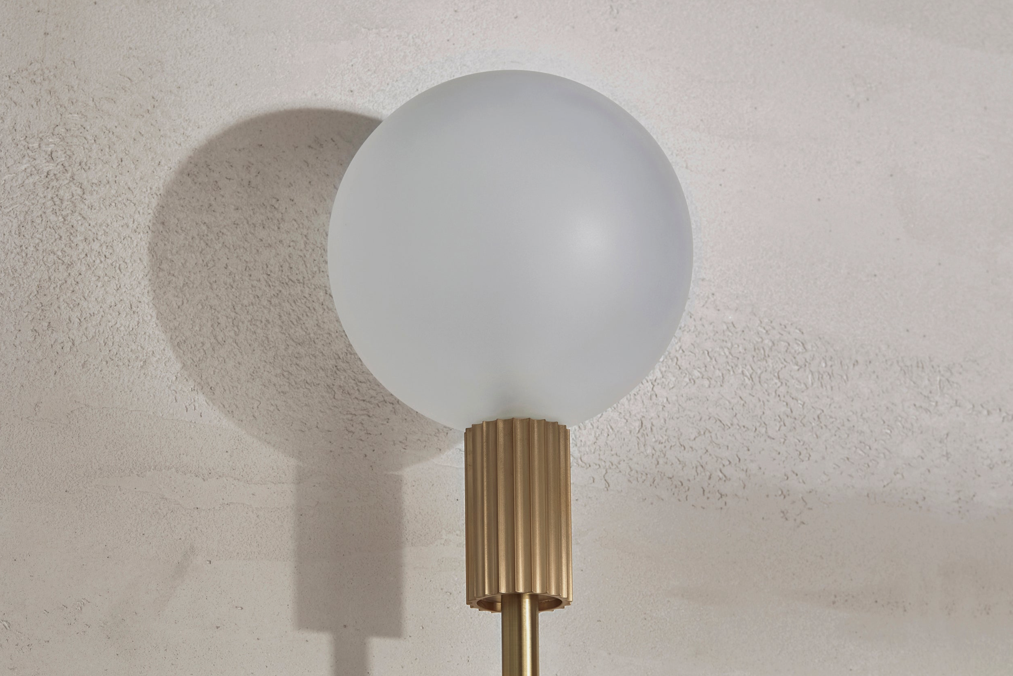 Wall-mounted indoor lamp with a white globe shade and gold base against a textured wall.