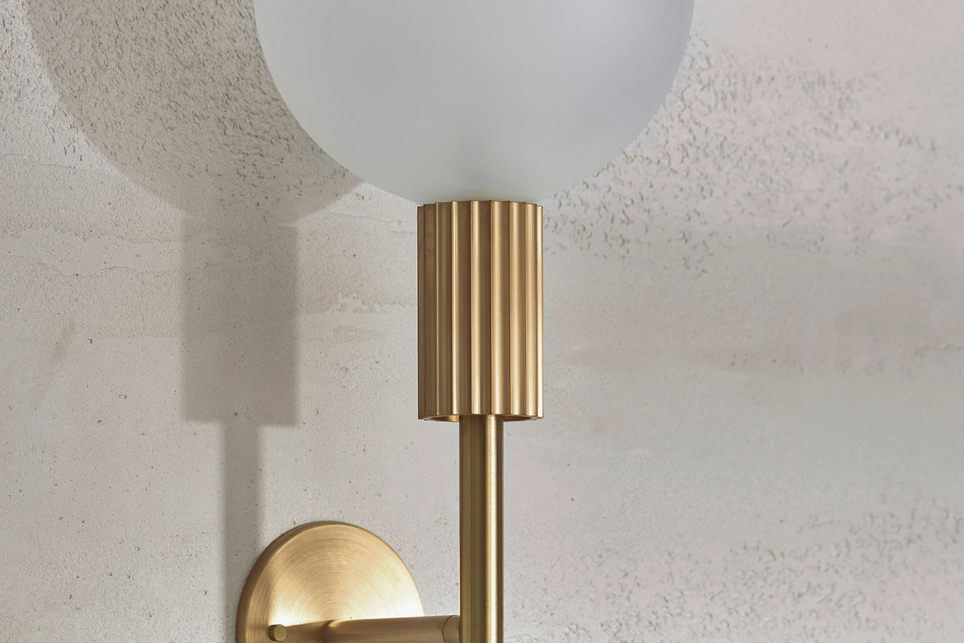 Wall-mounted indoor light fixture with gold finish and white glass shade on a textured wall.