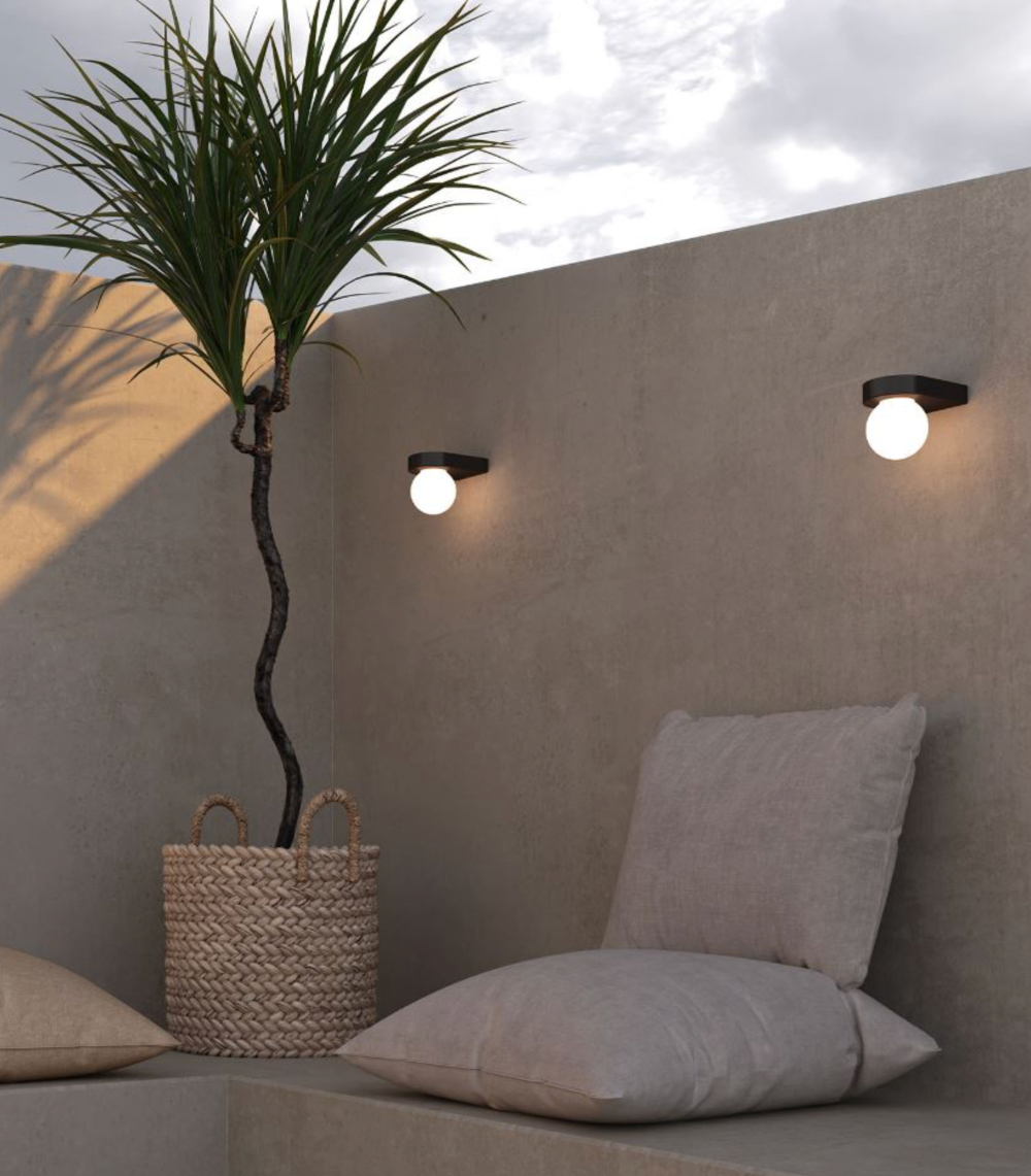 Outdoor seating area with a plant, pillows, and wall-mounted lights against a beige wall.