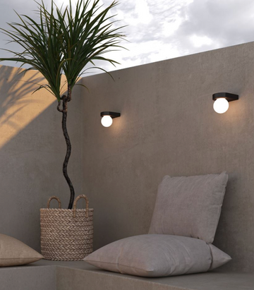 Outdoor seating area with a plant, pillows, and wall-mounted lights against a beige wall.