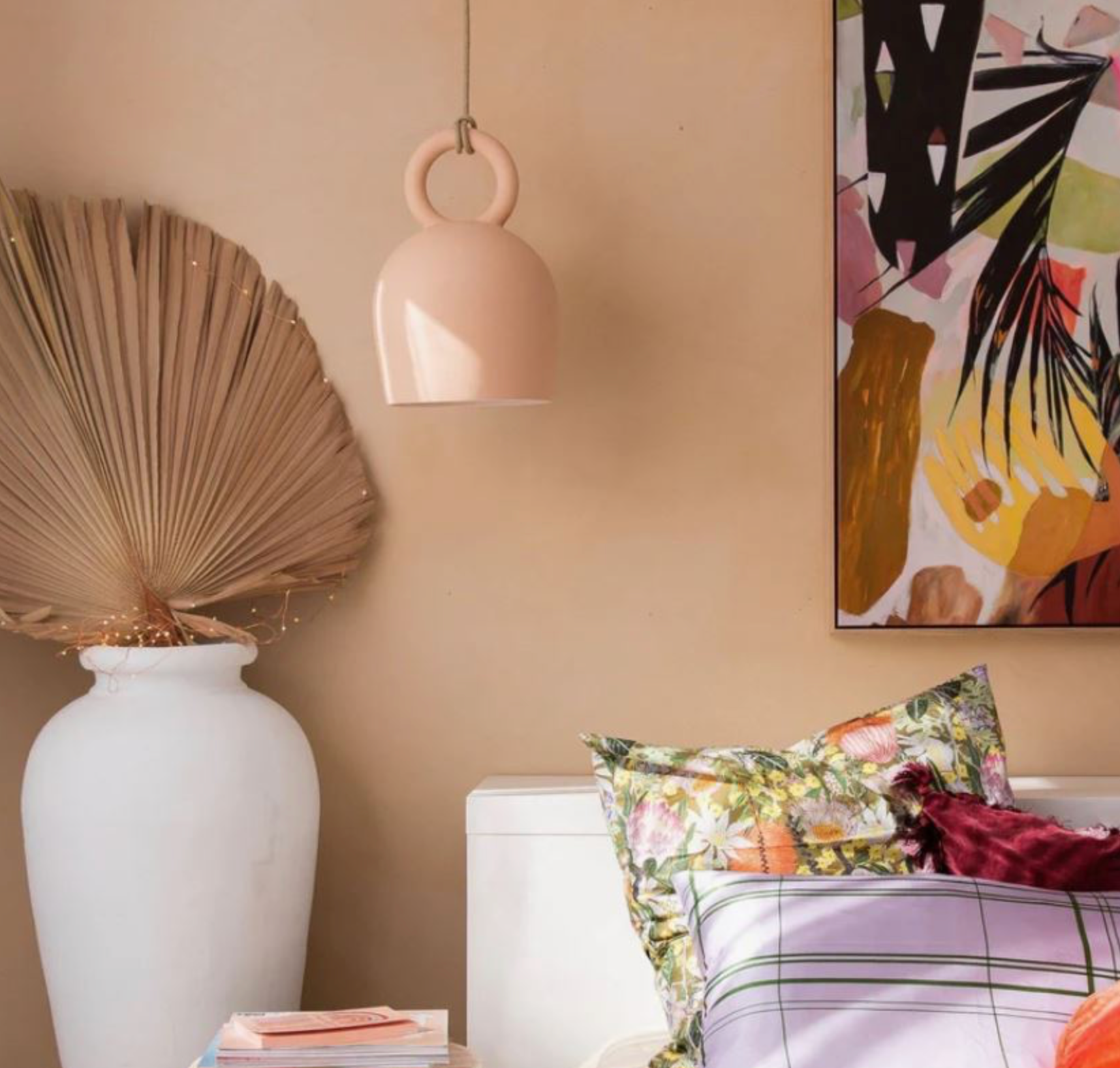 Decorative elements in a room including a white vase with dried palm leaves, a pink pendant light, and a colorful abstract painting.