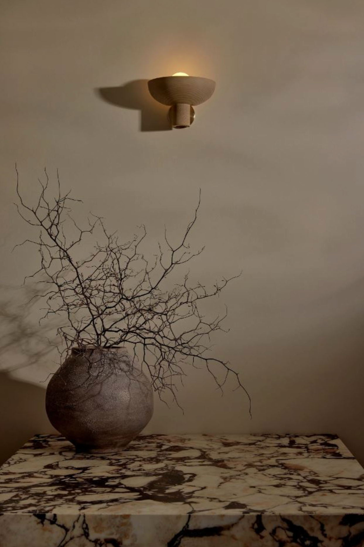 Decorative vase with branches on a marble surface under a wall-mounted light fixture.