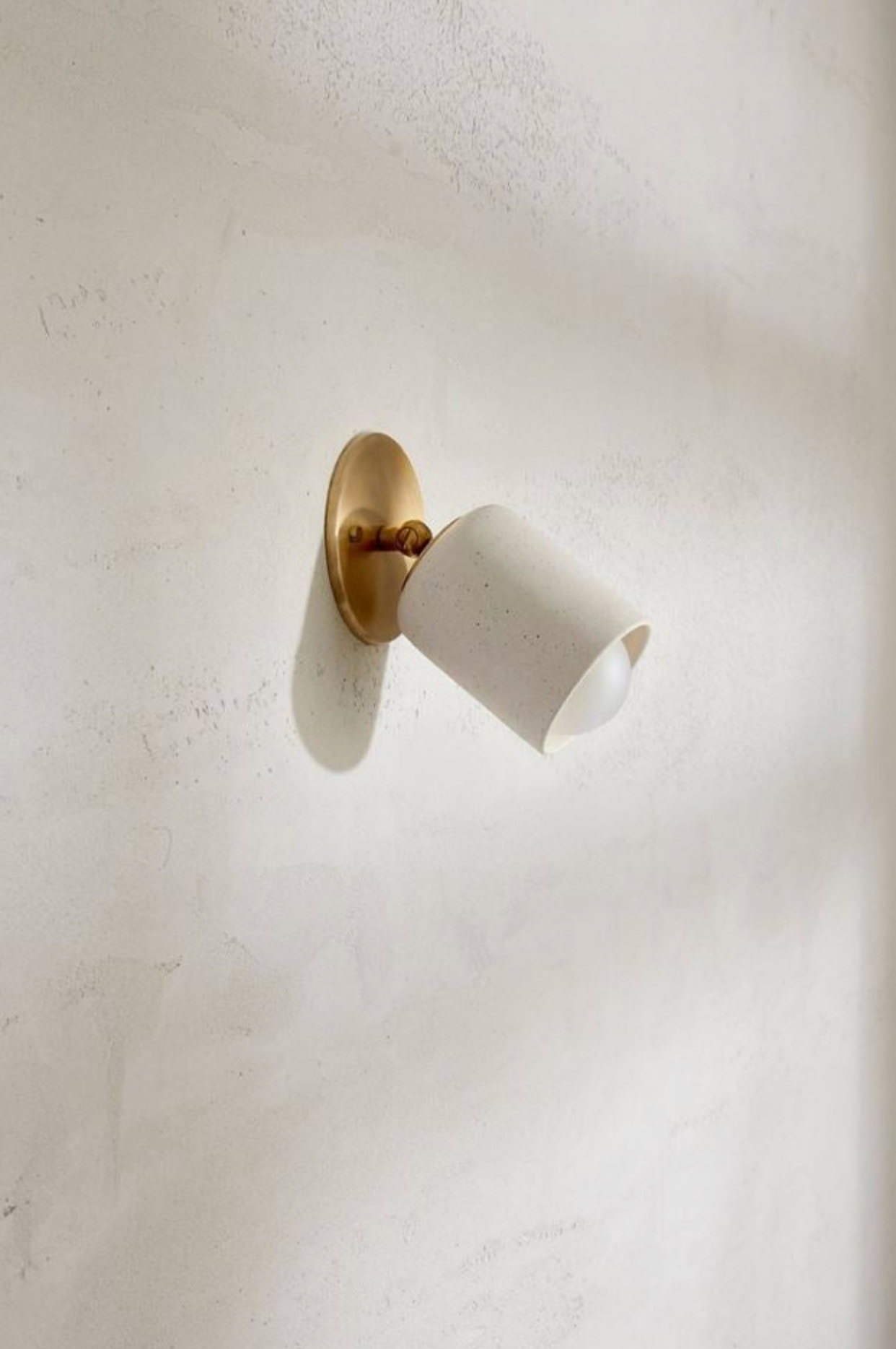 Wall-mounted light fixture with white shade and gold fixture on a plain wall.