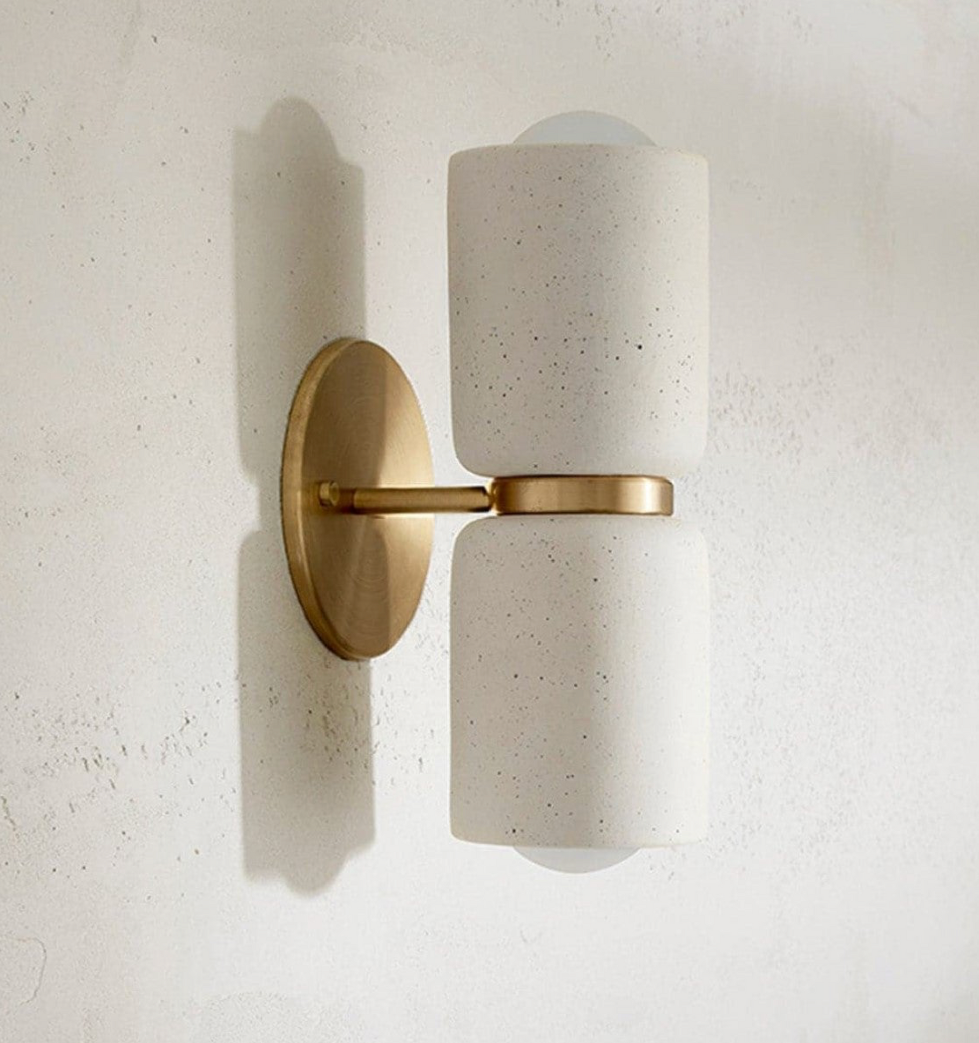 Wall-mounted ceramic light fixture with white cylindrical shade and gold metal base on a textured wall.