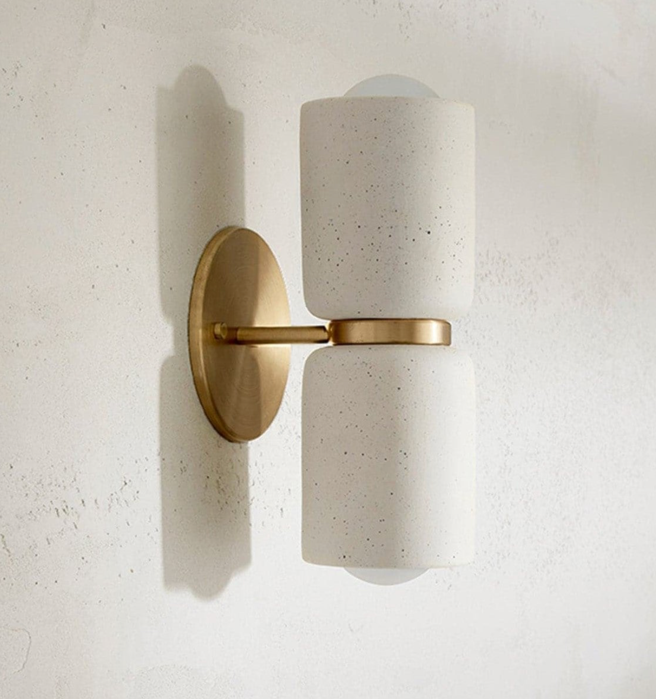 Wall-mounted ceramic light fixture with white cylindrical shade and gold metal base on a textured wall.