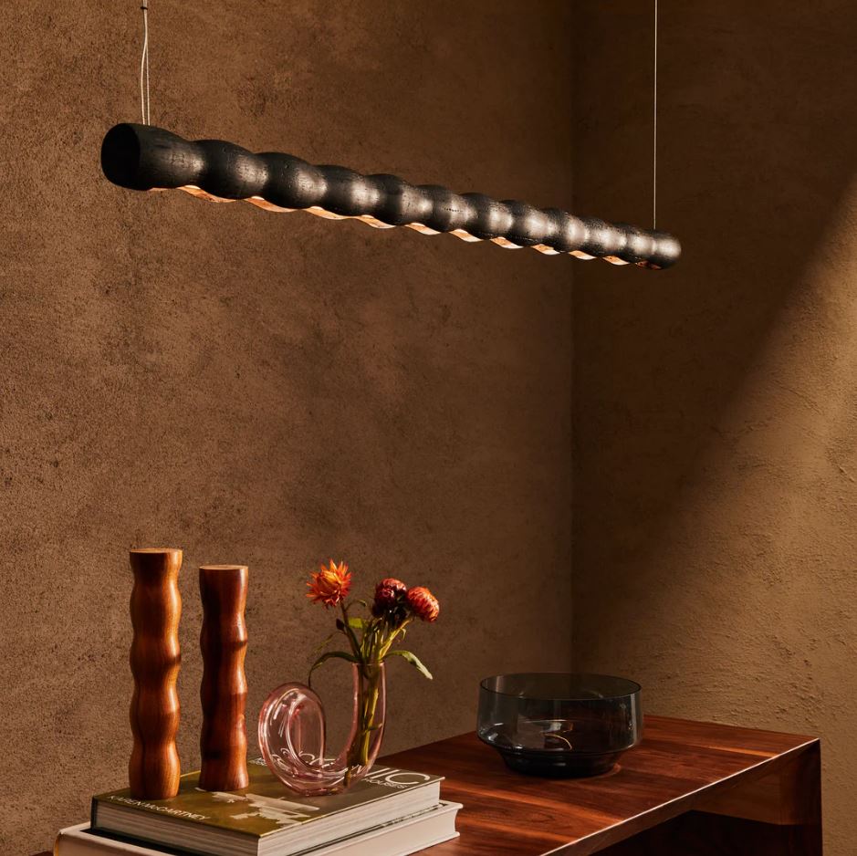 Modern wooden linear pendant light fixture hanging above a wooden table with decorative items.