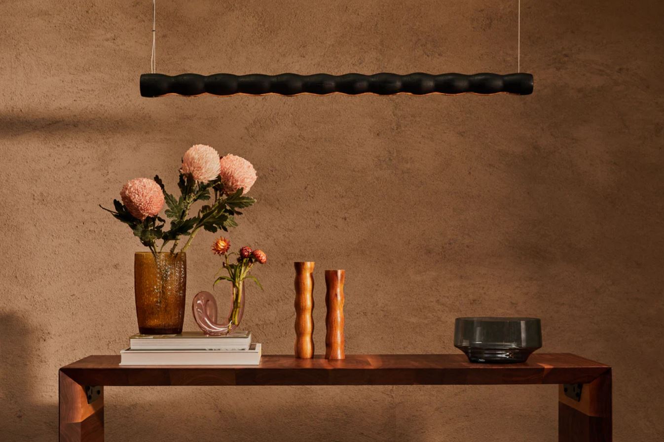 Decorative wood linear pendant light hanging over a wooden table against a brown wall with a black lamp.