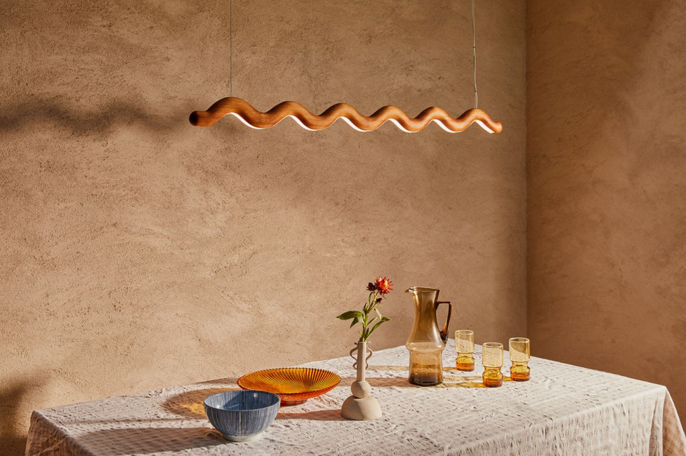Dining table with decorative items against a beige wall with a wavy wood linear pendant light fixture.