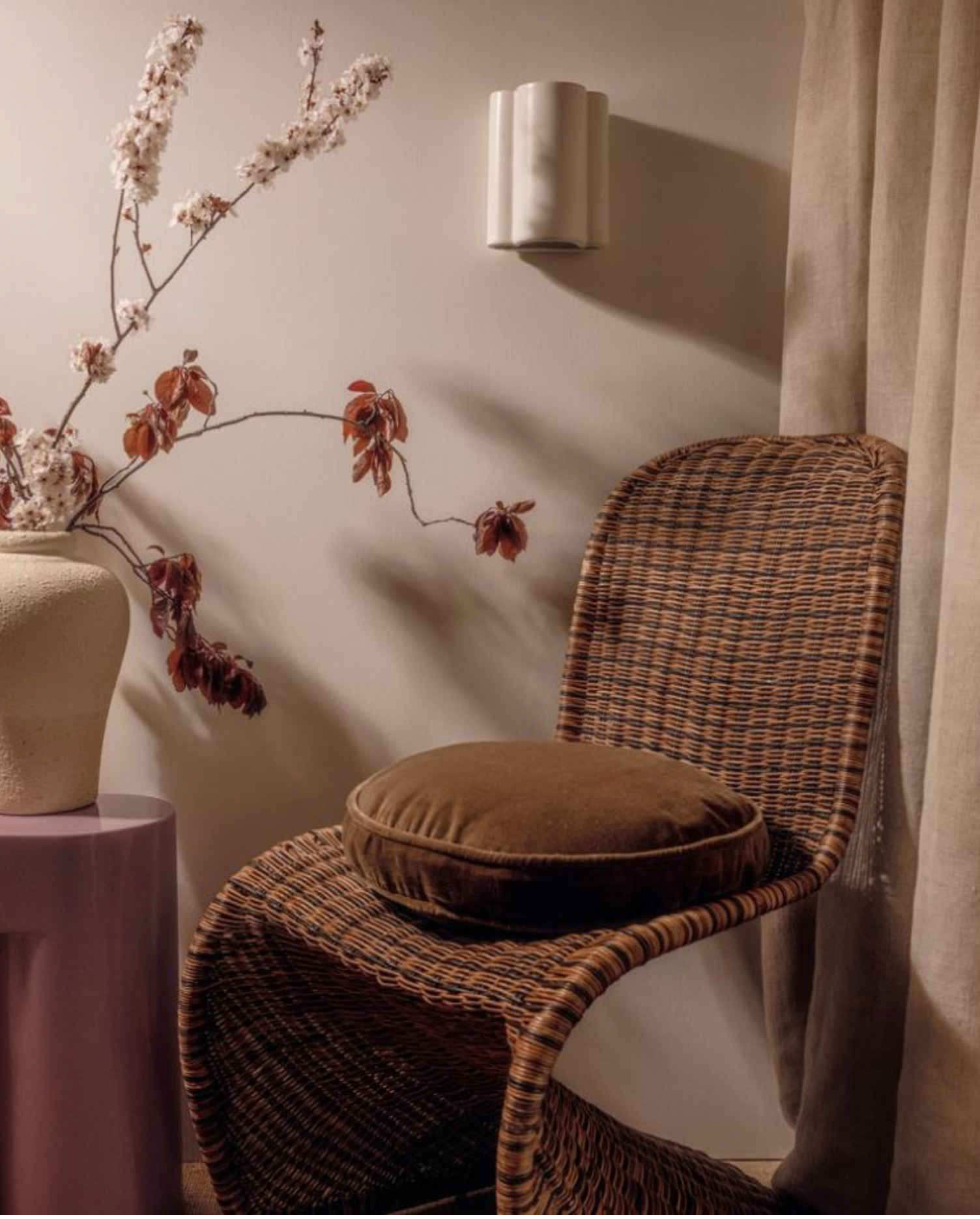 Wicker chair with a cushion next to a vase with flowers on a pink table against a neutral wall.