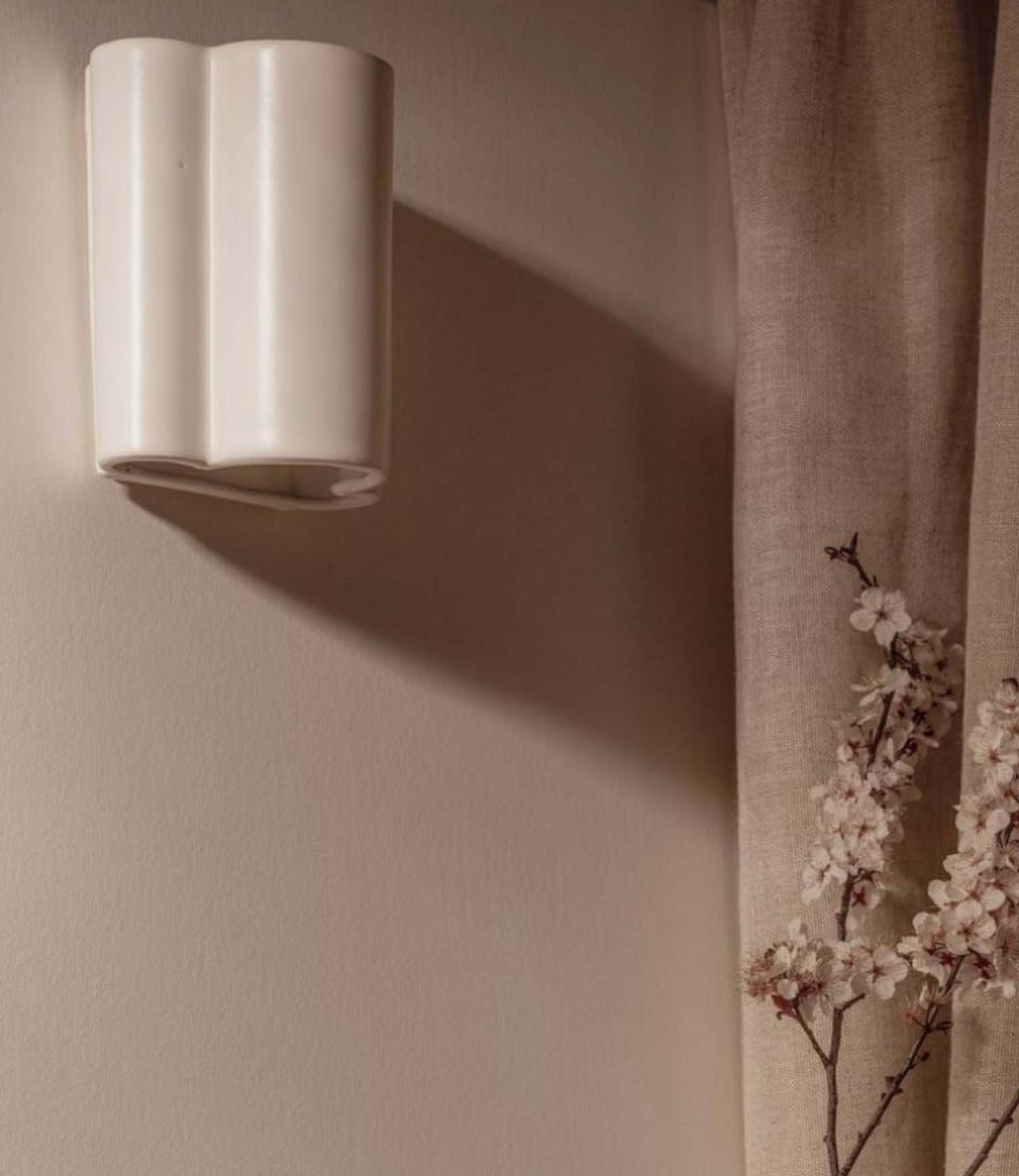 White wall-mounted ceramic light on a beige wall with a soft shadow, next to a curtain and floral branch.