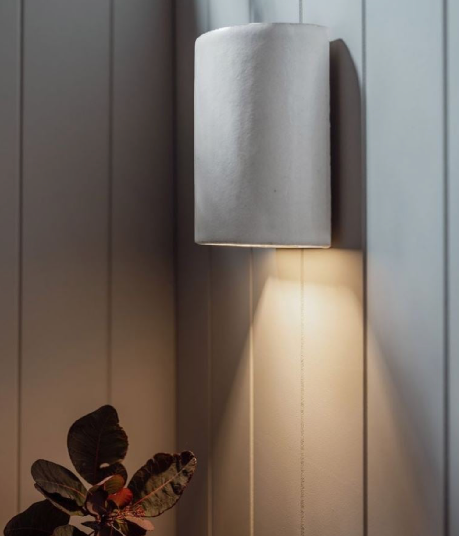 Cylindrical ceramic wall light fixture on a wooden panel wall with a plant in the foreground.