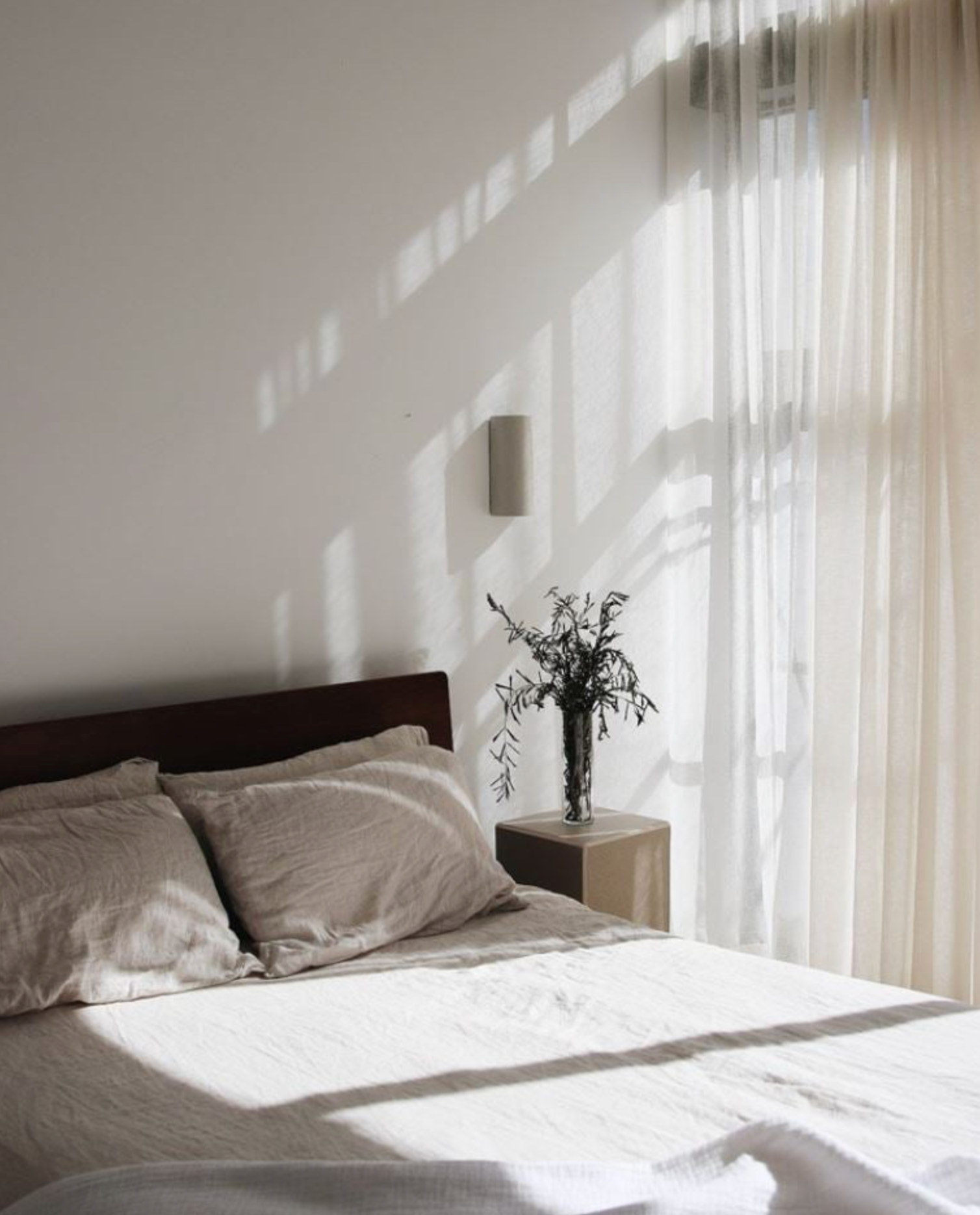 Bedroom with sunlight casting shadows on the interior wall light, featuring a bed and a small table with a vase.