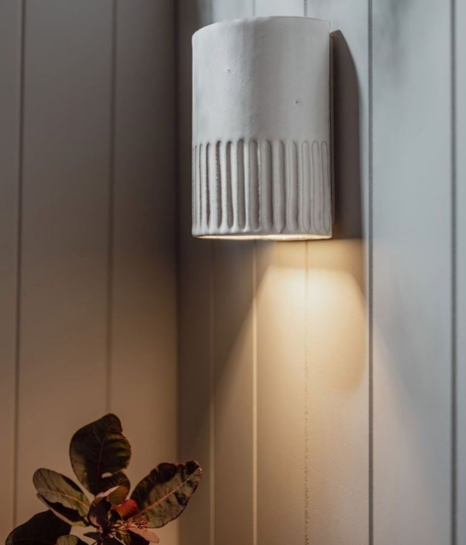 Ceramic wall light fixture on a white paneled wall with a plant in the foreground.